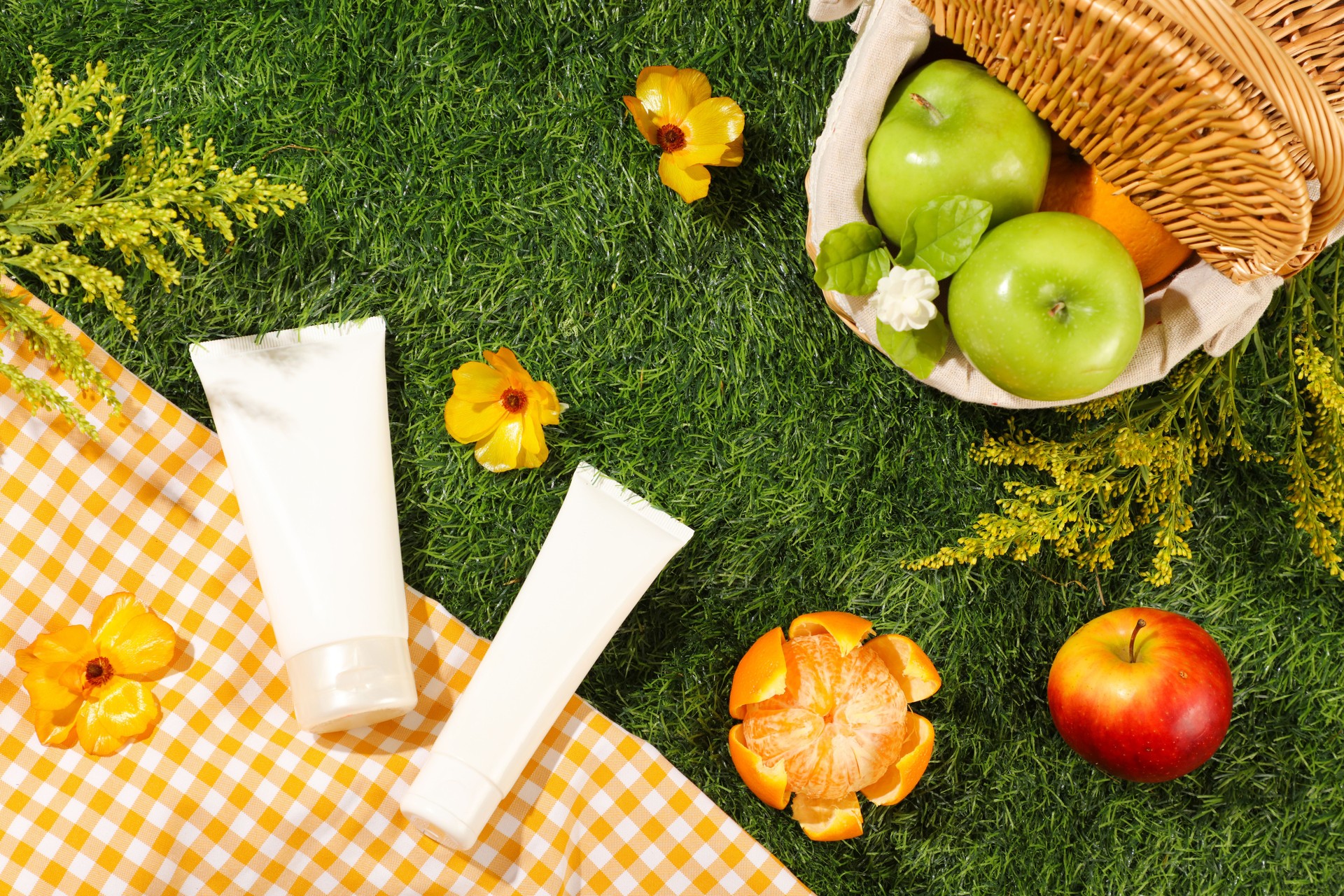 Top view of bamboo basket full with organic fruits on green grass background with checkered fabric and yellow flowers. Two cosmetic tubes unlabeled mockup for design packaging